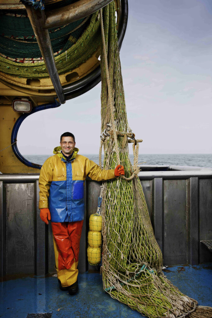 Pescatore in cerata gialla e blu accanto alla rete da pesca sul ponte di un peschereccio
